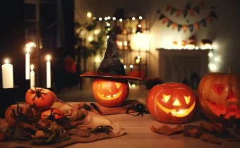 Halloweenské dekorace v podobě halloweenských dýní na jídelním stole. Vedle dýní hoří svíčky a v pozadí je krb, nad kterým jsou vyvěšeny malé vlaječky v oranžové a černé barvě.