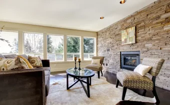 Útulný obývací pokoj s kamenným obkladem kolem krbu, pohovkou a křesly, doplněný velkými okny pro přirozené osvětlení.