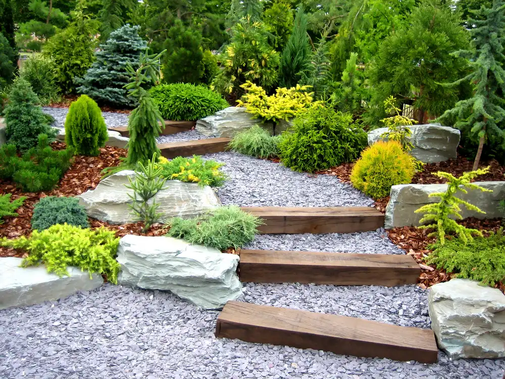 Zahradní cestička z šedé štěrkové drti s krokvy a obklopená různými druhy zelených rostlin a velkými kameny.