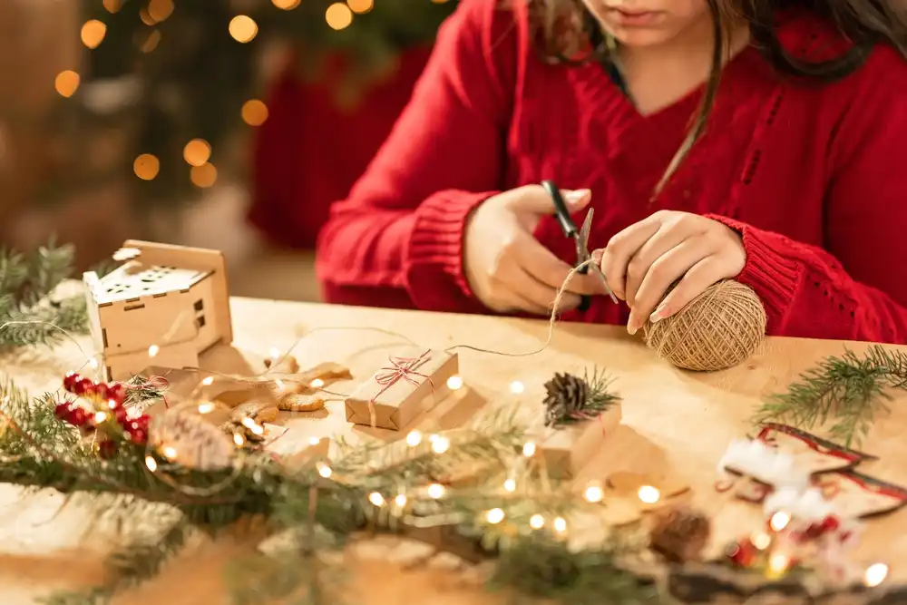 Dívka v červeném svetru tvoří vánoční dekorace, stříhá provázek a má před sebou vánoční světýlka a výzdobu, což naznačuje tvorbu DIY vánočních ozdob.