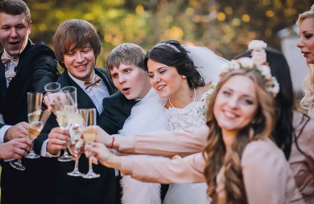 Skupina lidí oslavující na svatbě, drží si sklenice s šampaňským a připíjí, v popředí je družička s květinovým věncem na hlavě a hosté v elegantních oblecích.