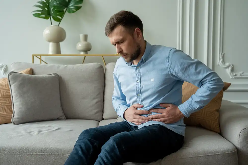 Muž sedí na pohovce a svírá si břicho, což může být známkou břišní bolesti nebo zácpy.