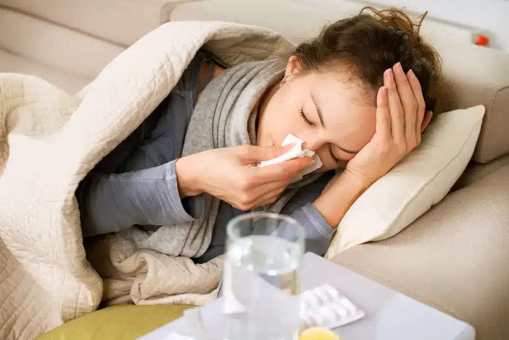 Žena ležící na gauči s horečkou, otírá si nos papírovým kapesníkem, obklopena léky a sklenicí vody, vypadá unaveně a nemocně.