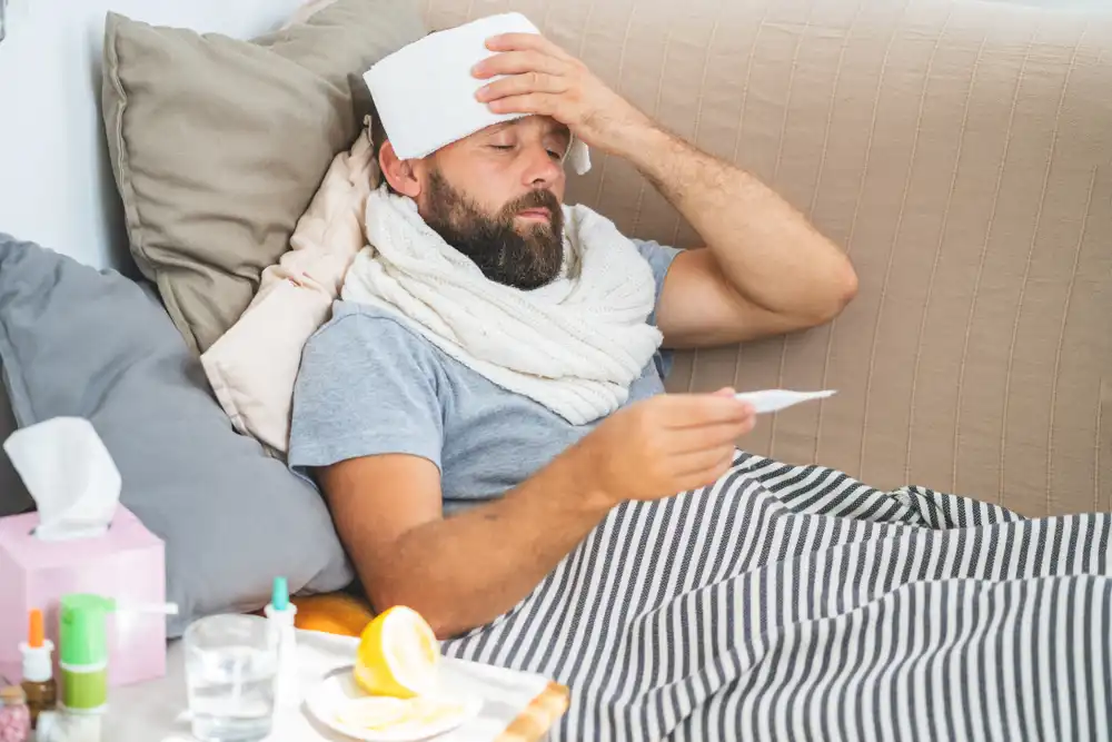 Muž sedí v posteli s ručníkem na hlavě, teploměrem a léky kolem sebe, naznačuje horečku a nemoc.
