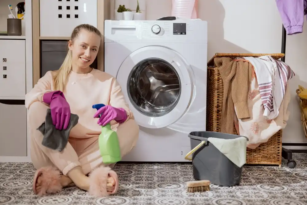 Usměvavá žena sedí na podlaze s čistícími prostředky a fialovými rukavicemi u otevřené pračky, připravená k čištění.