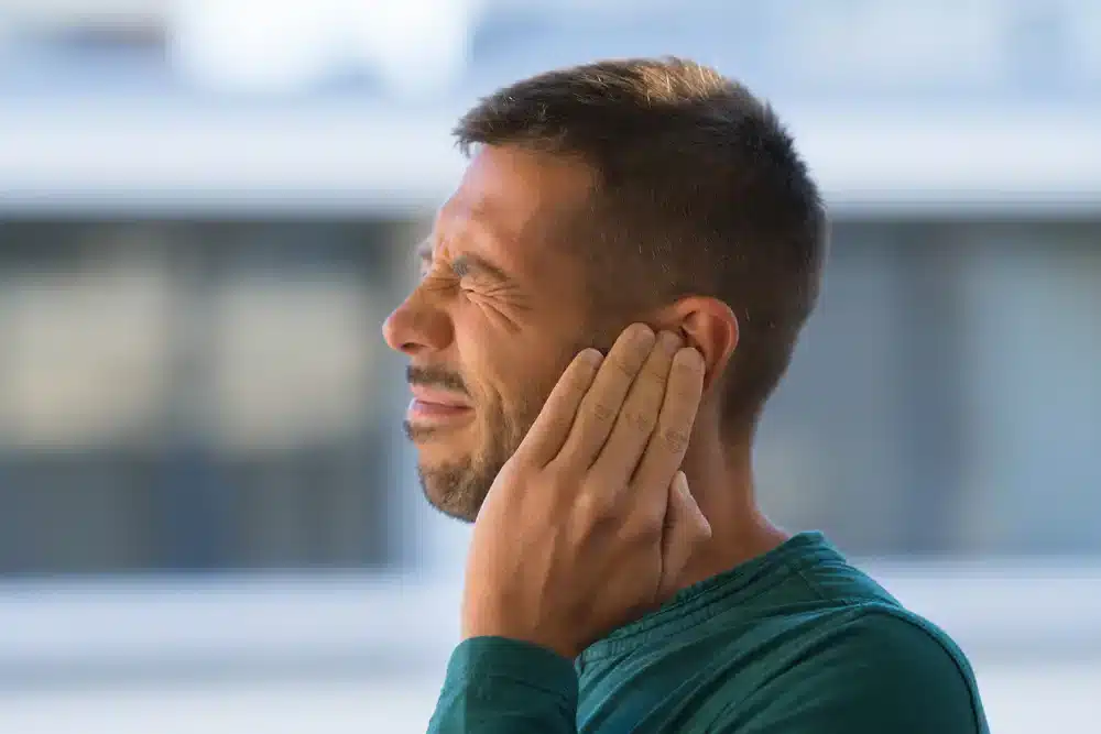 Muž v zeleném tričku si drží ucho a mračí se, což může být známka bolesti nebo nepohodlí způsobené zalehlým uchem.