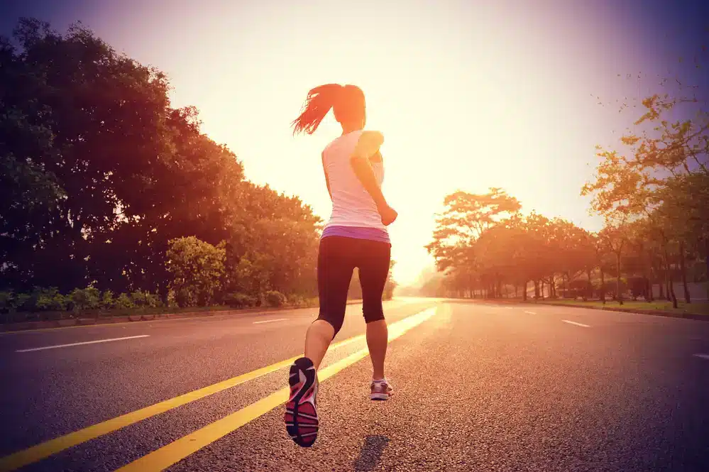 Žena běžící na prázdné silnici za svítání nebo soumraku, s výhledem na stromy a čistou oblohu. Je zezadu, oblečená v bílém tričku a černých legínách, což ztělesňuje pocit svobody a odhodlání při běhu.