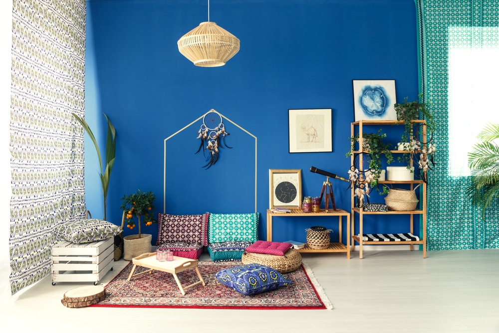 Interiér v boho stylu s výraznou modrou stěnou, vzorovanými textiliemi, ratanovým lustr, a dekorativními prvky jako jsou polštáře, rostliny, a obrazy.
