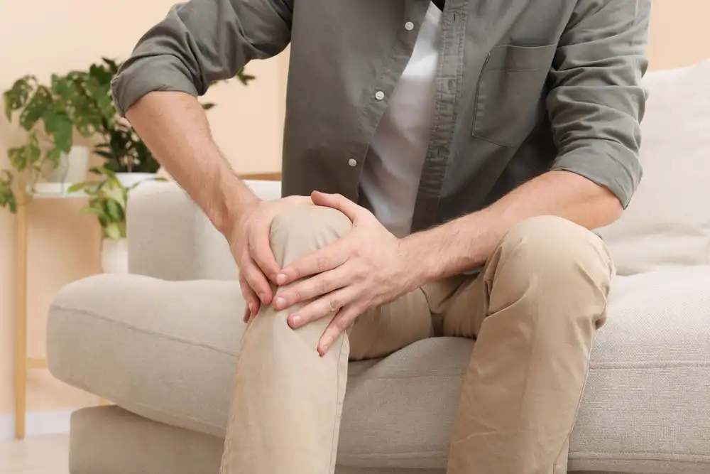 Muž sedící na pohovce drží rukou koleno, vypadá, že ho bolí.
