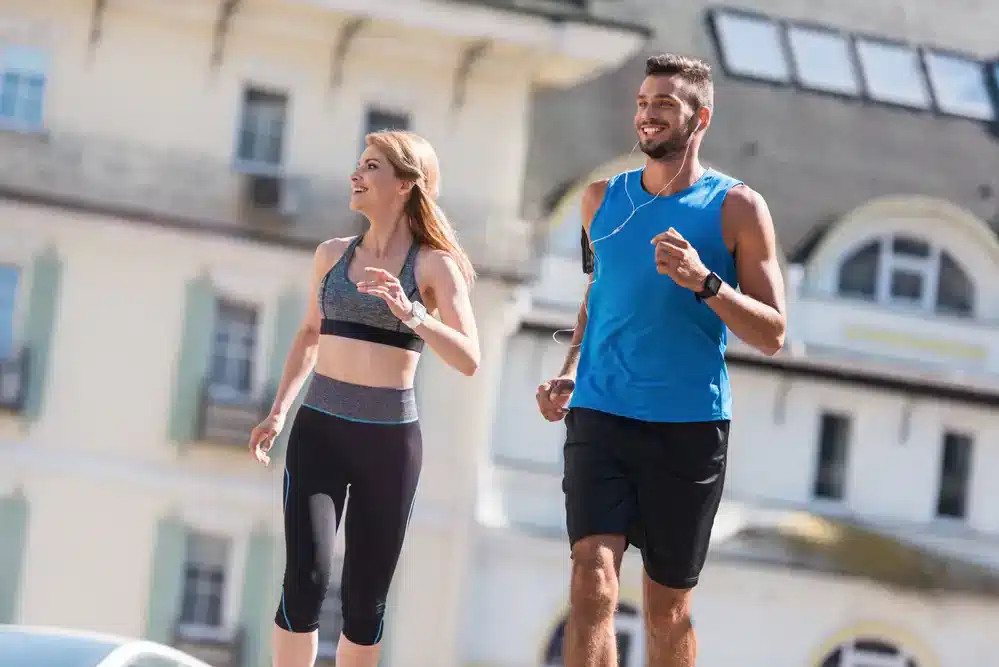 Muž a žena v sportovním oblečení běhají vedle sebe na městském chodníku. Muž má na sobě modré tričko a černé kraťasy, žena nosí šedý sportovní podprsenku a černé legíny. Oba vypadají šťastně a energeticky, což ilustruje radost z běhu.