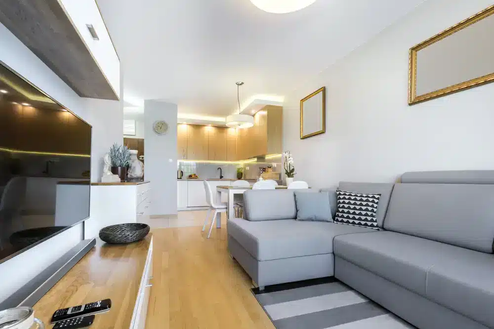 Světlý obývací prostor s kuchyňským koutem, bílé skříňky a dřevěným stolem.