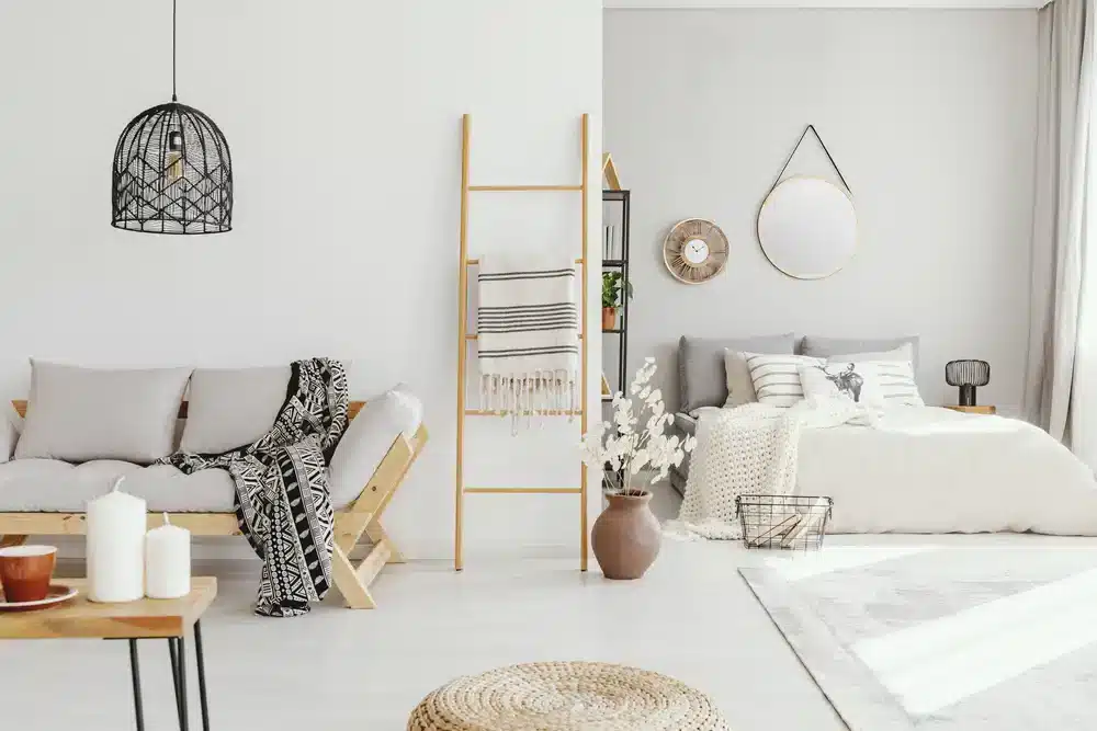 Obrázek zobrazuje obývací pokoj s minimalistickým designem. Bílé stěny, jednoduchý dřevěný nábytek a neutrální barevné tóny vytvářejí pocit prostoru v malém bytě. Detaily jako vzorovaná deka a keramická váza přidávají texturu a osobnost.