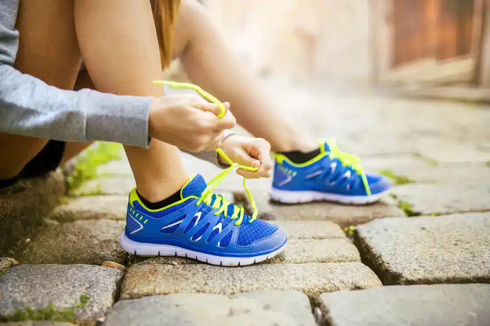 Detailní záběr na nohy ženy sedící na kamenném schodišti, zavazující si žluté tkaničky na modrých sportovních botách, připravujíc se na běh.