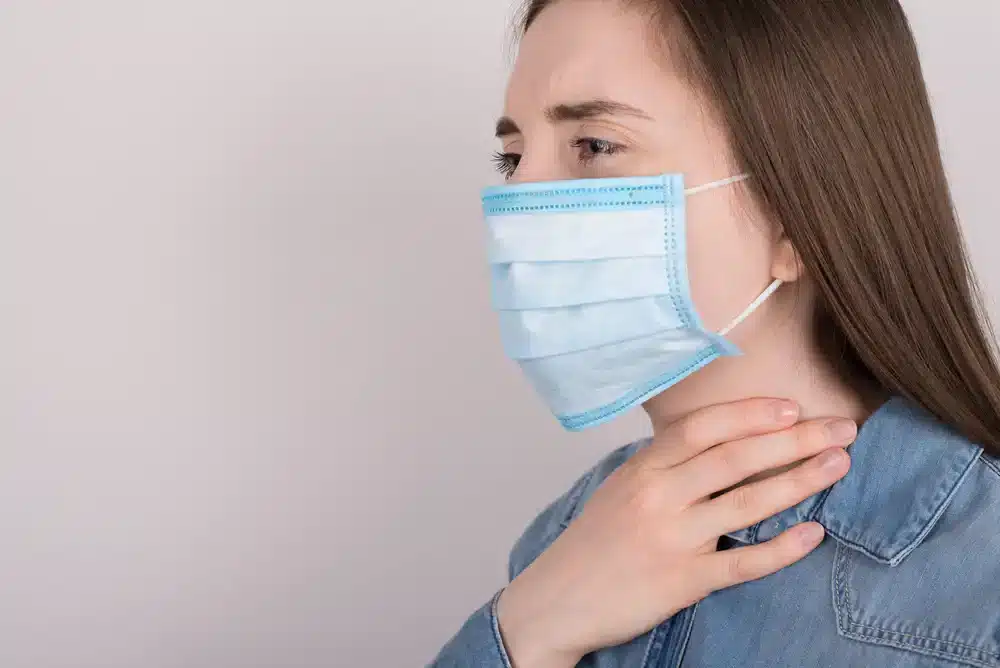 Žena s modrou ochrannou maskou na obličeji sahá si na hrdlo, známka možného bolestivého hrdla nebo obtíží s dýcháním.