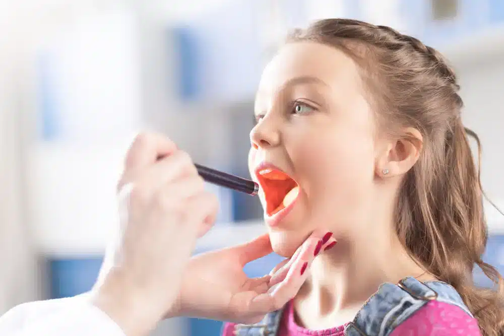 Malá dívka otevírá ústa, zatímco lékař ji vyšetřuje hrdlo, což je běžná praxe při diagnostice respiračních onemocnění jako je zánět průdušek.