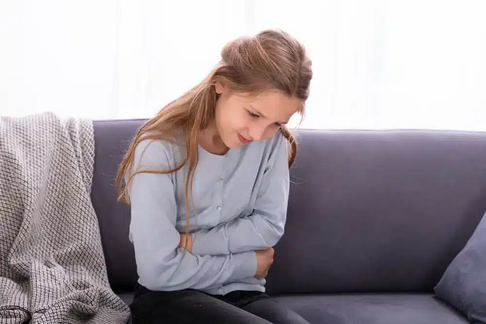 Dívka sedící na pohovce drží ruce na břiše, což může naznačovat bolest břicha nebo nevolnost.