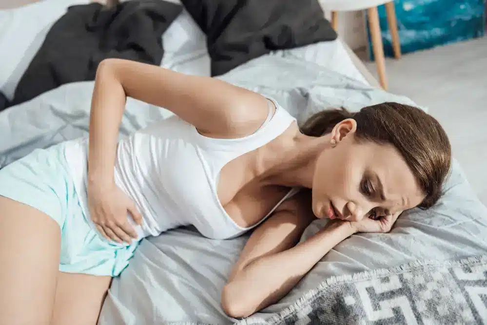 Žena ležící na boku v posteli s bolestivým výrazem v obličeji a rukou na břiše symbolizuje problémy s menstruačním cyklem.