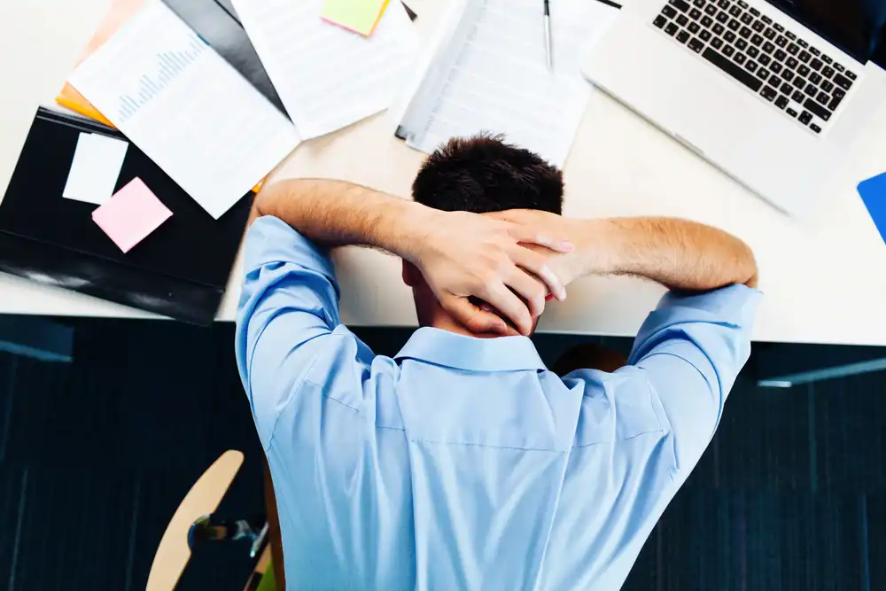 Muž v modré košili relaxuje v kanceláři s hlavou opřenou do rukou, což může pomoci snížit stres a tím i hladinu kortizolu.