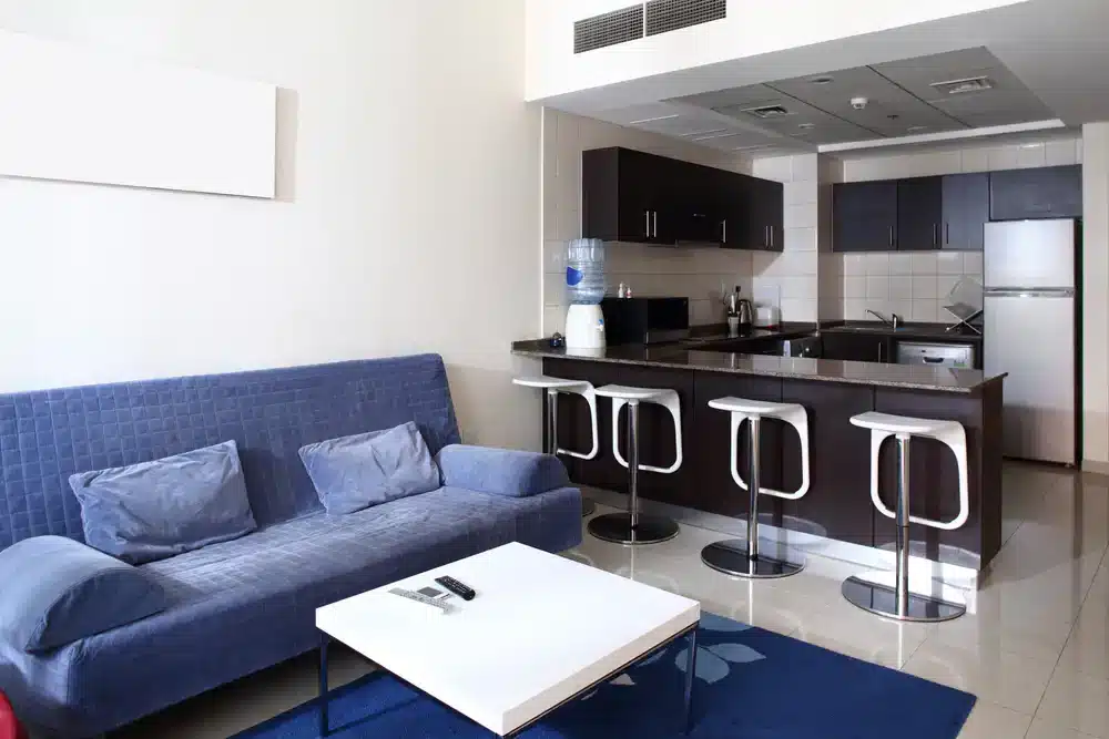 Funkční rozložení malého bytu s modrou pohovkou a kuchyňským ostrůvkem sloužícím jako jídelní stůl, což je praktické řešení pro 2+kk byt.