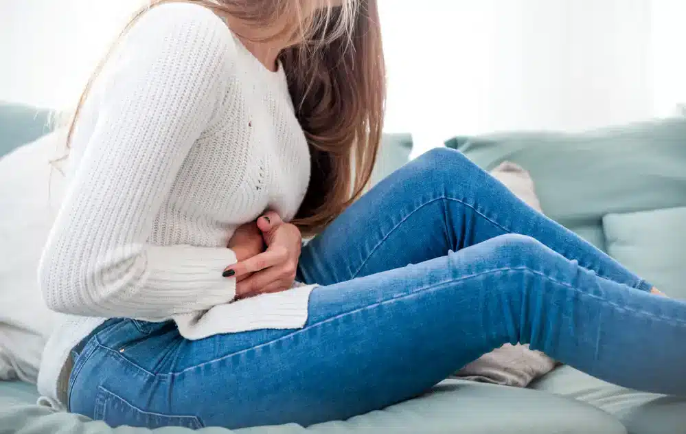 Zdrcená žena sedící na posteli se svírá za břicho, což ukazuje na intenzivní menstruační bolesti.