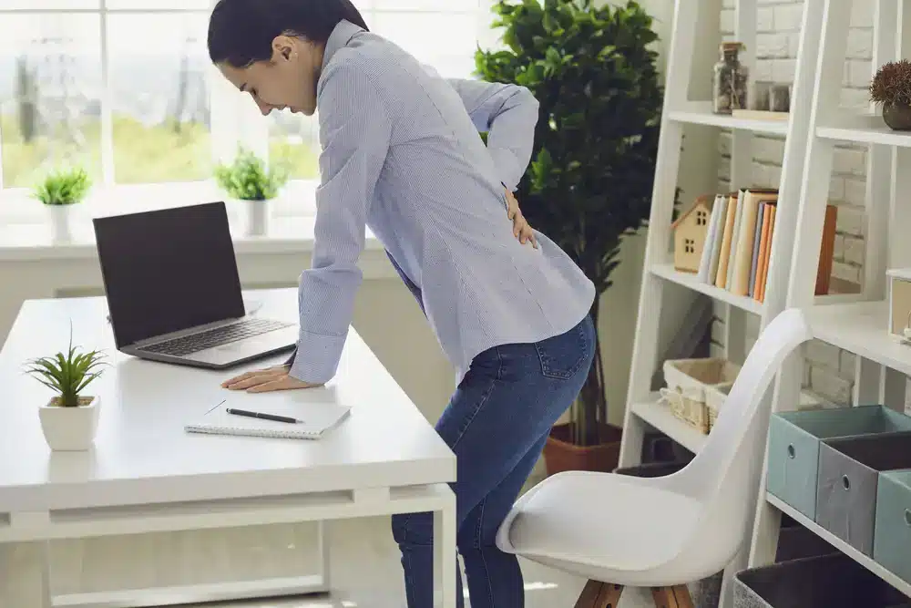 Pracující žena v kanceláři se sklání před notebookem a drží se za záda, což naznačuje možný ischias nebo zánět sedacího nervu.