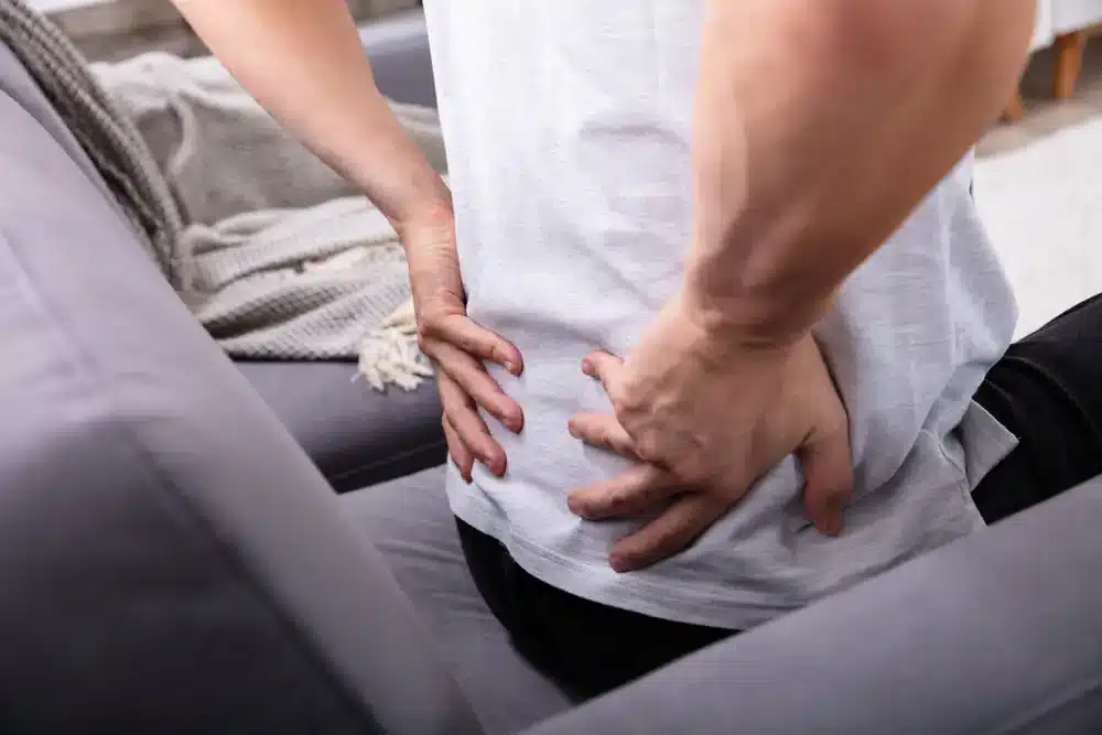 Muž sedící na pohovce a opatrně se drží dolní části zad, což může ukazovat na ischias nebo zánět sedacího nervu.