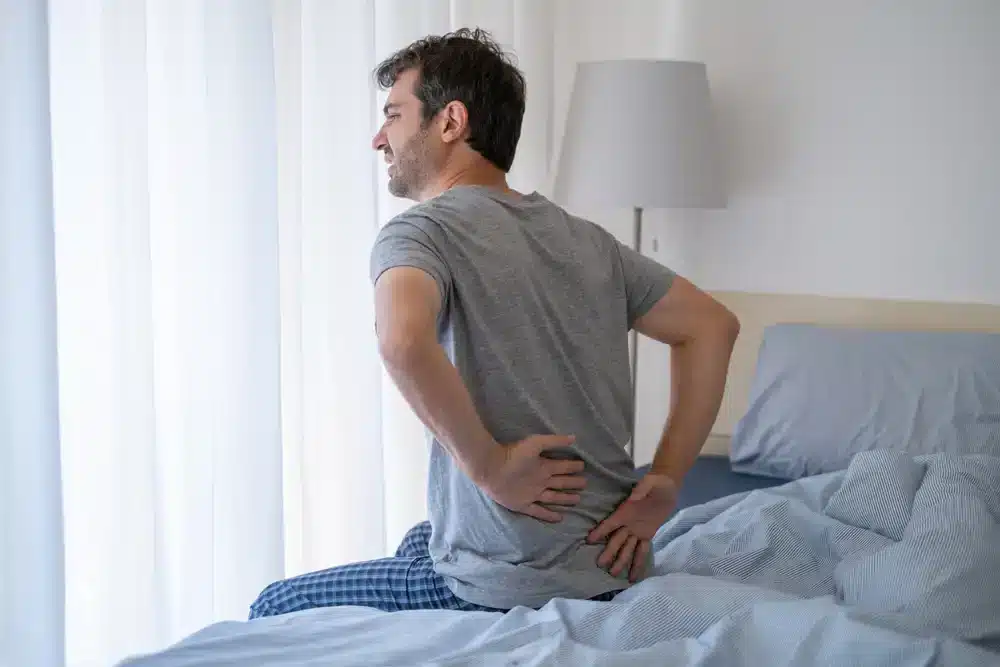 Muž sedící na posteli a držící se za bedra, vyjadřuje bolest, která může být příznakem ischiase nebo zánětu sedacího nervu.