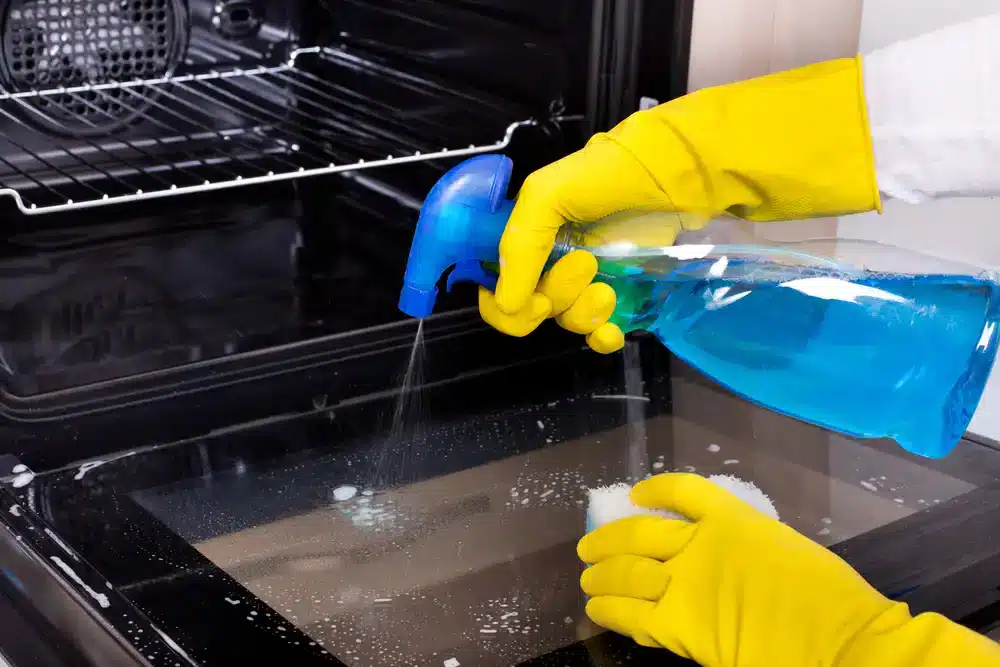 Aplikace čistícího prostředku na sklo trouby s použitím rozprašovače, metoda pro snadné a účinné čištění trouby.