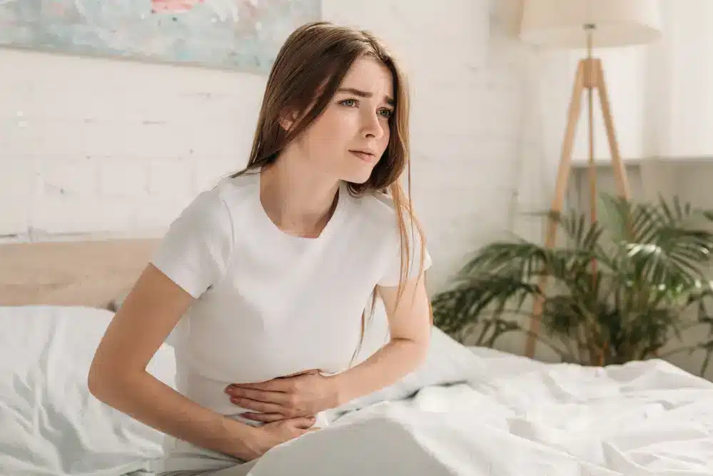 Mladá žena sedí na posteli s bolestivým výrazom a drží si břicho.